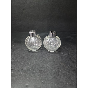 May include: Two clear glass salt and pepper shakers with silver-colored tops. The shakers have a round, ribbed design and are sitting on a dark surface. The tops have small holes for dispensing the contents.