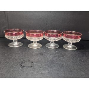 May include: Four vintage clear glass dessert dishes with a ruby red rim and a decorative pattern. Each dish has a short stem and a round base. The dishes are arranged in a row against a dark background.
