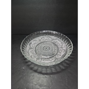 May include: A clear glass serving plate with a decorative floral and geometric pattern. The plate has a scalloped edge and a raised center. This vintage dish is suitable for serving appetizers or desserts.