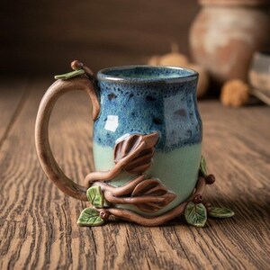 May include: A ceramic mug featuring a two-tone blue and light green glaze. The exterior is decorated with sculpted brown branches, small green leaves, and tiny berries that wrap around the base and extend up the side to the handle.