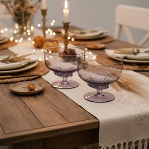 May include: Two light purple glass dessert bowls with a wide, shallow basin and a short, sturdy pedestal base. The bowls feature a smooth, translucent finish and are positioned on a wooden table, showcasing a simple, functional design for serving.