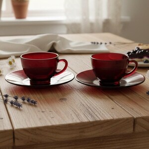 May include: Two matching deep red glass teacups with rounded bowls and curved handles, each resting on a coordinating circular glass saucer. The translucent red glassware features a smooth finish and a consistent, vibrant crimson colour throughout.