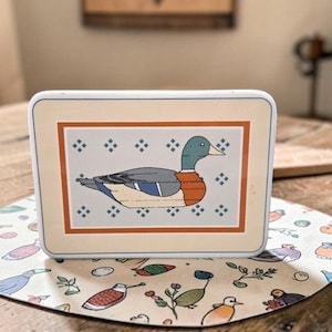 May include: A rectangular, off-white trinket box with a duck illustration. The duck is depicted with a green head, gray body, and orange chest. The box has a brown border and is set on a patterned mat with birds and floral designs.