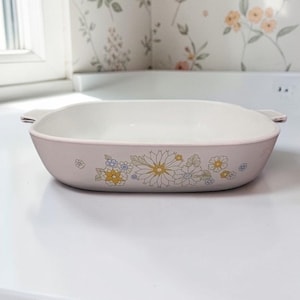 Pode incluir: Uma assadeira retangular rosa claro com interior branco e design floral. A assadeira tem duas pequenas alças e é decorada com flores amarelas, azuis e brancas. Está sobre uma superfície branca.