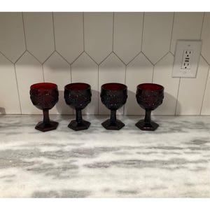 May include: Four ruby red wine glasses with ornate floral designs. The glasses have a hexagonal base and a short stem. The glasses are arranged in a row on a marble countertop.