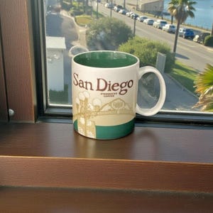 Puede incluir: Una taza de cerámica blanca con interior y base verdes, con las palabras "San Diego" en letras marrones. La taza también muestra un gráfico de un puente y el logotipo de Starbucks Coffee.