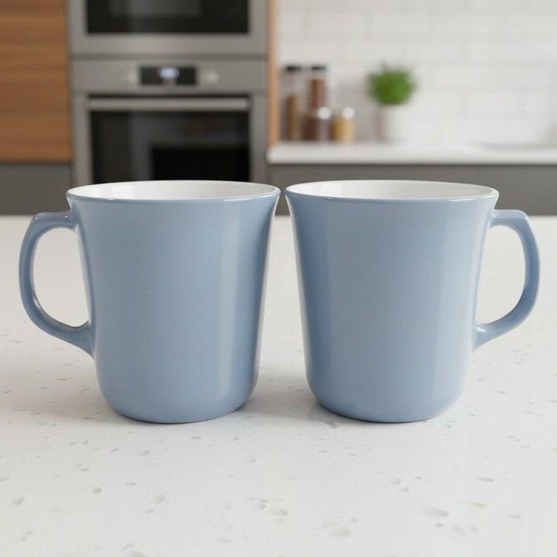 Two light blue ceramic coffee mugs with white interiors stand side by side on a white countertop against a blurred kitchen backdrop.
