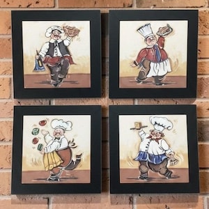 May include: Four framed ceramic tiles with cartoon chefs in different poses. Each chef wears a white hat and apron, holding food items such as bread, bottles, and vegetables. The frames are black, and the background is a brick wall.