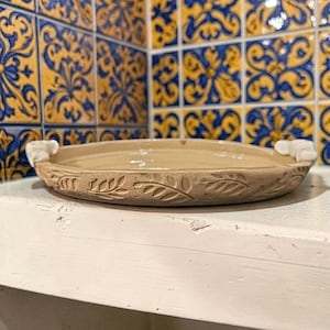 May include: A beige ceramic soap dish with a leaf pattern around the edge and small white handles. The dish is oval-shaped and sits on a white surface. The background features blue and yellow patterned tiles.