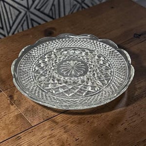 May include: A clear, vintage glass serving dish with a scalloped edge. The dish is divided into sections, with a central starburst design and a diamond-patterned border. The dish is likely used for serving appetizers or snacks.