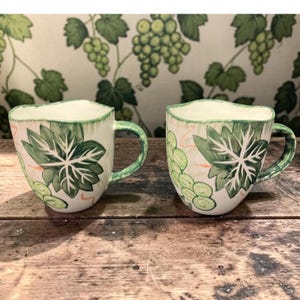 May include: Two ceramic mugs with a cream-colored base, featuring green grape leaf and vine designs. The rims and handles are painted green. The mugs are set on a wooden surface.