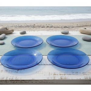 May include: Four blue glass plates arranged on a white wooden table with a light blue placemat. The plates have a textured rim and a smooth center. The background shows a beach scene with the ocean and sand.