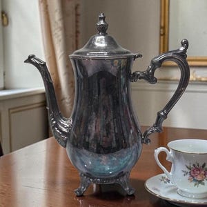 Op de afbeelding: Een zilverkleurige koffiepot met een gebogen tuit en een sierlijke handgreep. De pot heeft een deksel met een decoratieve knop en staat op drie kleine voetjes. Een kop en schotel met bloemmotief staan ernaast.