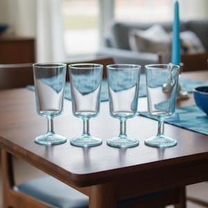 Op de afbeelding: Vier heldere wijnglazen met een lichtblauwe tint, gerangschikt op een houten tafel. De glazen hebben een klassiek ontwerp met een afgeronde voet en een slanke steel. De tafel is gedekt met een blauwe loper en een blauwe kaars.
