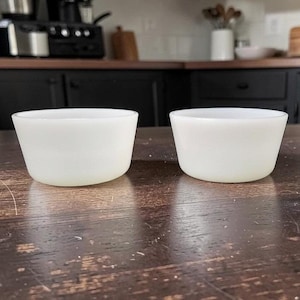 May include: Two small, off-white glass bowls with a slightly flared shape. The bowls are set on a dark wooden surface, with a kitchen setting in the background. The bowls are likely for serving or food preparation.
