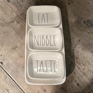 Puede incluir: Una bandeja de cerámica blanca con tres compartimentos rectangulares. Cada compartimento tiene una palabra en letras grises: "EAT", "NIBBLE" y "TASTE". La bandeja está sobre una superficie de madera.