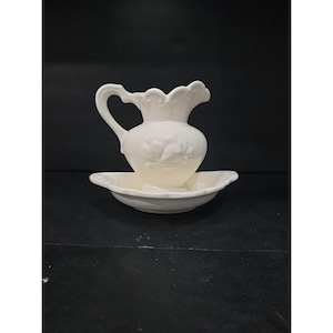 May include: A white ceramic pitcher with a curved handle and a scalloped rim, resting on a matching shallow basin. The pitcher features a floral relief design. The set is likely for decorative use or serving water.