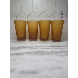 Puede incluir: Cuatro vasos de color ámbar con un diseño floral. Los vasos son altos y cilíndricos, con un aspecto ligeramente esmerilado. Están dispuestos en fila sobre una encimera de mármol, con un salpicadero de azulejos blancos al fondo.