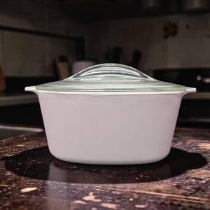 May include: A light pink, vintage casserole dish with a clear glass lid. The dish has a rounded shape with small handles on either side. The lid has a curved handle. The dish is sitting on a dark wooden surface.