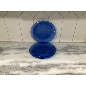 May include: Two round, translucent cobalt blue glass plates. One plate is positioned vertically, standing upright, while the second plate is placed horizontally in front of it, creating a stacked appearance with a smooth, reflective surface.