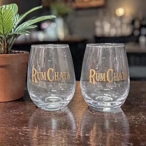 Puede incluir: Dos vasos transparentes sin tallo con las palabras "Rum Chata" en letras doradas. Los vasos están sobre una superficie de madera oscura. Una planta en maceta es visible en el fondo.
