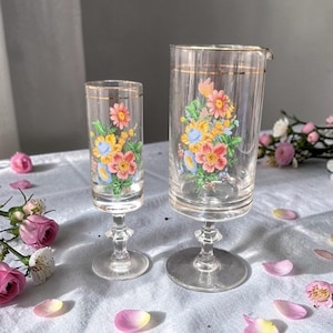 May include: Two vintage clear glass cocktail glasses with gold rims and floral designs. The glasses feature a stem and base. The floral design includes pink, yellow, and blue flowers. The glasses are on a white surface with flower petals.