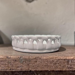 Op de afbeelding: Een kleine, ronde, witte keramische kom met een gevlochten ontwerp rond de rand. De kom staat op een rustiek houten oppervlak. Het ontwerp van de kom heeft een reeks kleine, gelijkmatig verdeelde openingen.