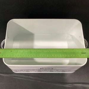 An open white bread box with metal handles viewed from above, measured with a green ruler across the interior.