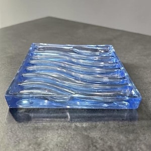 Può includere: Un portasapone quadrato blu trasparente con un motivo a onde. Il portasapone è in vetro e ha una forma rettangolare. Il design a onde aggiunge un elemento decorativo all'oggetto funzionale.