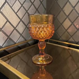 May include: Amber-colored vintage goblet with a diamond pattern on the bowl. The stem and base are also amber, and the glass reflects light. The goblet is likely made of pressed glass and is a decorative item.