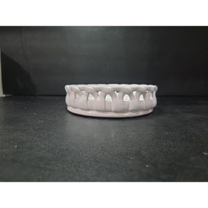 May include: A small, round, ceramic basket with a woven design. The basket is painted in a soft, muted lavender color. The basket has a series of openings around the rim, adding to its decorative appeal. The basket is suitable for holding small items.