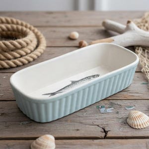 May include: A rectangular ceramic baking dish with a light blue exterior and a white interior. The dish features a ribbed outer texture and a black and white illustration of a single fish printed on the bottom of the interior basin.