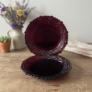Op de afbeelding: Twee ronde borden van donkerrood geperst glas. Elk bord heeft een geschulpte rand en een decoratief randpatroon bestaande uit reliëfovalen en kleine stippen. Het midden van elk bord is glad en reflecterend en toont de donkerrode kleur.