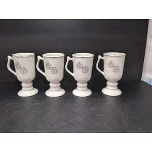 May include: Set of four white pedestal mugs with gold trim and a decorative emblem. Each mug has a handle and a footed base. The mugs are arranged in a row against a dark background.
