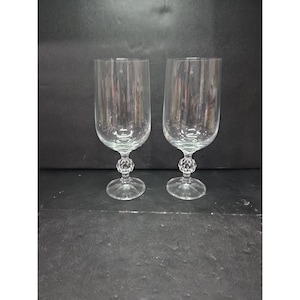 May include: Two clear, stemmed water goblets. The glasses have a rounded bowl shape and a decorative, faceted ball on the stem. The base is a flat, circular disc. The goblets are made of clear glass and are set against a dark background.
