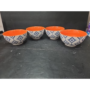 May include: Four ceramic bowls with orange interiors and off-white exteriors. The exterior features a repeating blue geometric pattern. The bowls are arranged on a dark surface.