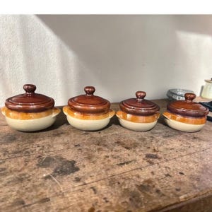 May include: Four small, ceramic lidded pots with a classic design. The pots have a cream-colored base, a band of orange, and a dark brown lid and handle. The pots are arranged in a row on a wooden surface.