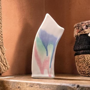 Könnte beinhalten: Eine weiße Keramikvase mit geschwungenem Design. Die Vase hat pastellblaue, grüne und rosa Farbtropfen. Die Vase steht auf einem Holzregal, mit einem geflochtenen Korb und einem Seil im Hintergrund.