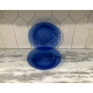 May include: Two round, translucent cobalt blue glass plates stacked vertically. The plates feature a smooth, glossy finish with a simple, circular design. The glass is uniform in color, allowing light to pass through the material, creating a vibrant blue hue.