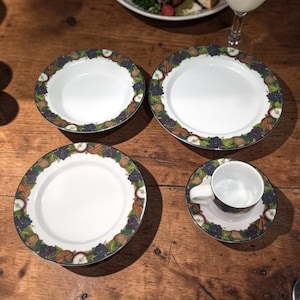 Puede incluir: Un juego de vajilla de cerámica blanca con un borde decorativo con un patrón de frutas y hojas en tonos verdes, morados y marrones. El juego incluye platos, un cuenco y una taza de té con un platillo, todo dispuesto sobre una superficie de madera.