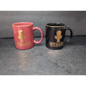 May include: Two ceramic mugs. One is burgundy with gold trim and a gold graphic that says "Nutcracker Ballet". The other is black with gold trim and a gold graphic that says "Nutcracker Ballet". Both mugs have a handle.