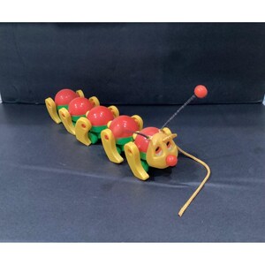 May include: A pull-along toy caterpillar featuring a yellow head with a red nose and a long black antenna with a red ball tip. The body consists of four segments with red spherical tops, green bases, and yellow legs, attached to a yellow pull string.