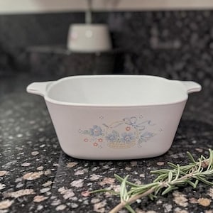 Pode incluir: Uma travessa branca, quadrada, com pegas. A travessa apresenta um desenho floral com flores azuis e uma cesta amarela. A travessa está sobre uma superfície escura e salpicada.