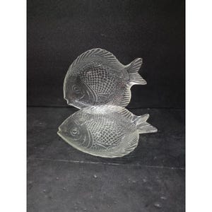 May include: Two clear glass fish-shaped dishes. The dishes have detailed scale patterns and fin designs. The fish are positioned one on top of the other, with the top fish slightly angled. The background is dark.