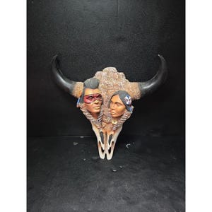 May include: A decorative buffalo skull with black horns and a detailed carving of two Native American faces. The skull is painted with earthy tones and features intricate details, creating a unique piece of art.