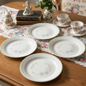 Puede incluir: Un juego de cuatro platos de cerámica blanca, cada uno con un delicado patrón de corona floral en tonos pastel en el centro y un borde verde fino. Los platos están dispuestos sobre una mesa de madera junto a dos tazas y platillos a juego.