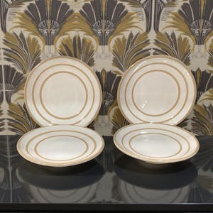May include: A set of four white ceramic plates with two concentric tan-colored rings around the rim. The plates are arranged on a dark surface, with a patterned wallpaper background in shades of gold, brown, and gray.