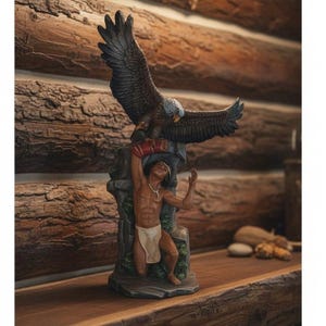 Puede incluir: Una escultura detallada que representa una figura arrodillada con un taparrabos blanco y un collar, extendiendo los brazos hacia arriba para sostener un gran águila de plumas oscuras con las alas extendidas. La base tiene textura de roca gris.