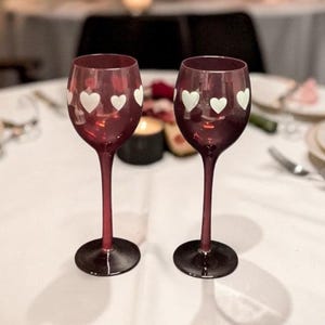 Può includere: Due calici di vino bordeaux con disegni a cuore bianchi. I calici hanno gambi lunghi e basi rotonde. Sono appoggiati su una tovaglia bianca, suggerendo un'occasione romantica o celebrativa. I calici sono vuoti.