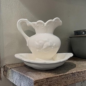 May include: A white ceramic jug with a decorative handle and a scalloped rim, sitting on a matching shallow dish. The jug and dish are displayed on a rustic wooden shelf, evoking a vintage feel.